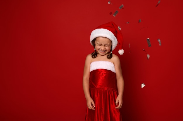 Mooi klein meisje in Santa carnaval kleding verheugt zich, poseert met gesloten ogen, schattige glimlach tegen rode achtergrond met vallende pailletten en confetti. Kerstmis, Nieuwjaarsviering concept, kopieer ruimte