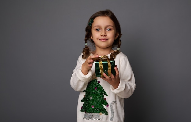 Mooi klein meisje in witte trui met een kerstboom, poseert tegen een grijze achtergrond, met een kerstcadeau in groen glitter inpakpapier en gouden lint, schattige glimlach die naar de camera kijkt