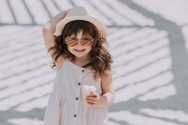 Mooi klein meisje in zonnebril witte jurk en hoed die ijs eet op het strand in de zomer