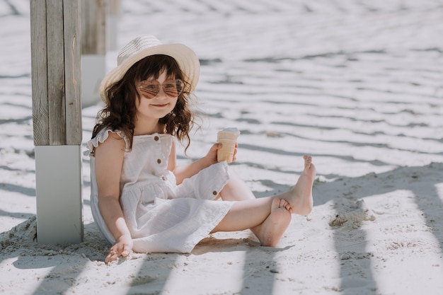 Mooi klein meisje in zonnebril witte jurk en hoed die ijs eet op het strand in de zomer