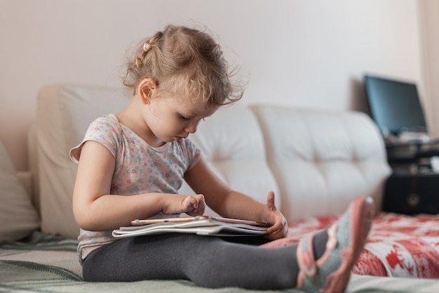 mooi klein meisje met een tablet, speelt en zit thuis op de bank. Afstandsonderwijs en een kind