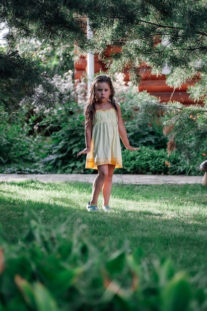 Mooi klein meisje met verbaasde ogen handen evenwijdig aan de grond staande onder dennentakken, gekleed in lichte zomerjurk en slippers in park overdag Geweldige achtergrond vol groen