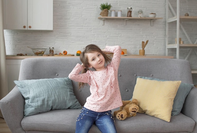 Mooi klein meisje speelt met haar eigen haar zittend op de bank tegen de achtergrond van het interieur