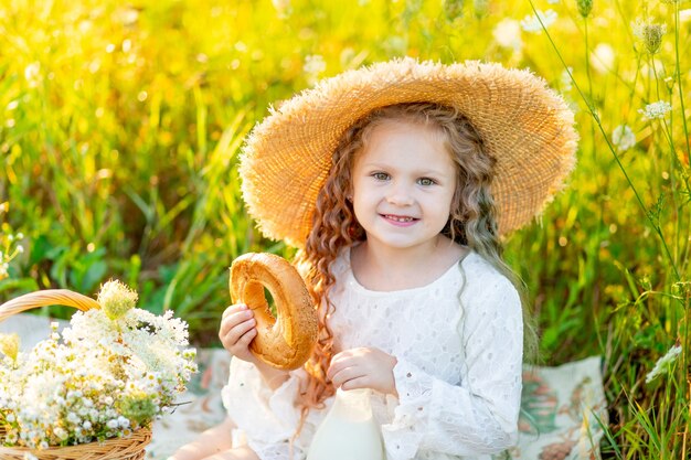 mooi klein meisje, zittend in een strooien hoed in een geel veld met wilde bloemen met een fles melk en een bagel