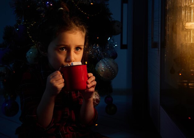 Mooi klein meisje zittend op de vloer bij de kerstboom en cacaodrank drinkend met marshmallows, haar gezicht verlicht door het licht van de oven met een duits stollenbrood op de bakplaat