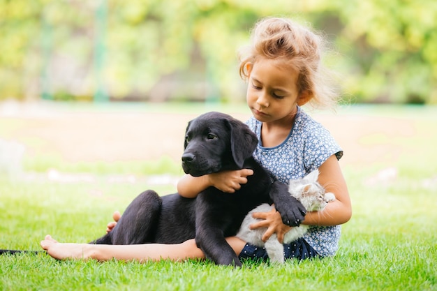Mooi klein meisje zittend op het gras met wit katje op haar knieën en omarmt zwarte puppy Meisje kijkend naar hond in haar armen Het verzorgen van huisdieren concept