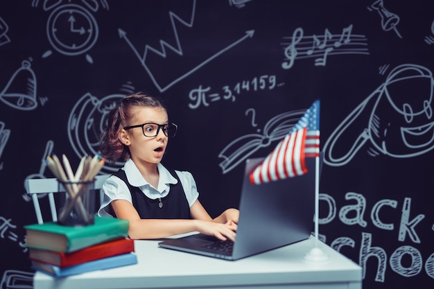 Mooi klein schoolmeisje bij bureau tegen zwarte achtergrond met de vlag van de v.s