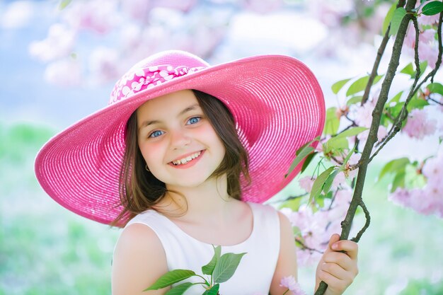 Mooi klein tienermeisje in roze modieuze hoed met lachend gezicht in de bloesem van de lentebloem op zonnige dag Lente tienerportret