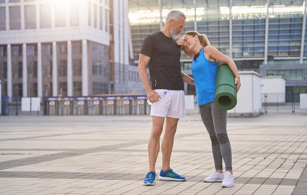 Mooi koppel van middelbare leeftijd in sportkleding die buiten staat met yogamat na ochtendtraining in