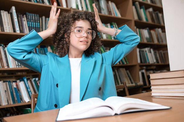 Mooi krullend meisje in glazen en blauwe pak zitten met boeken in de bibliotheek.