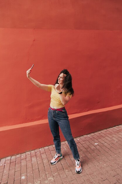 Mooi lachend model met afro krullen kapsel gekleed in zomer hipster kleding. Sexy zorgeloze vrouw poseren in de straat in de buurt van muur. Grappige en positieve vrouw die plezier heeft. Maakt selfie-foto's