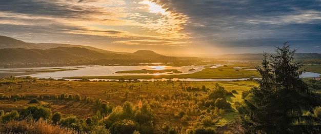 Mooi landschap met bergen en heuvels, rivier en bomen verlicht door de stralen van de rijzende zon door de wolken aan de hemel. screensaver, lay-out, mockup, natuurlijk concept.