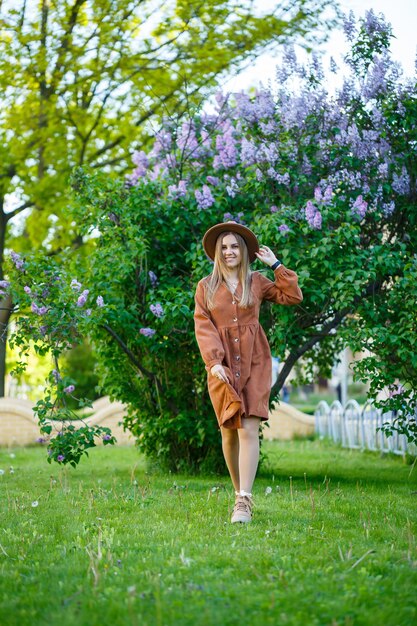 Mooi langharig meisje in een hoed op een achtergrond van lila bloemen. Ze heeft een Europese uitstraling met een glimlach op haar gezicht. Jonge vrouw die op een warme zonnige dag in een bloeiend park loopt