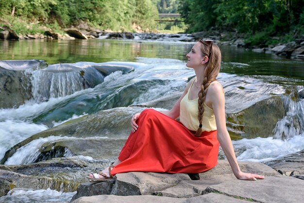 Mooi langharig meisje in een rode rok zittend op een rots. Zomer avond. Rust en genot van de natuur.