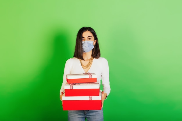 Mooi meisje dat beschermend gezichtsmasker draagt dat de dozen van giften houdt die over groene achtergrond worden geïsoleerd