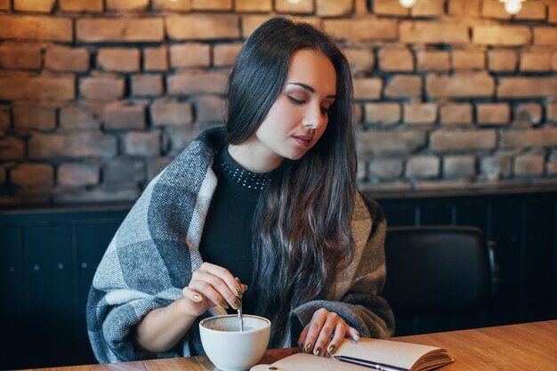 Mooi meisje drinkt koffie in een restaurant. Portret van een jonge dame met donker krullend haar die haar ogen droomachtig sluit met een kopje in haar handen. Mooi meisje zit in een café met een kopje koffie