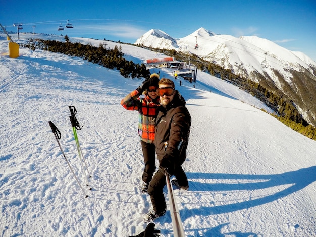 Mooi meisje en man die een lachende foto met selfiestick nemen op een berg bedekt met sneeuw.