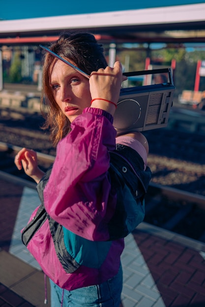 Foto mooi meisje gekleed in de stijl van de jaren 90 met draagbare radio-ontvanger in haar handen die zich voordeed op het perron van het treinstation