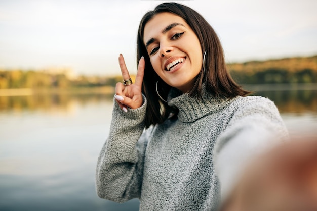 Mooi meisje glimlachend in het algemeen trui dragen zelfportret maken en vrede gebaar tonen Mooie gelukkige jonge vrouw selfie nemen op haar apparaat buiten tegen de natuur achtergrond