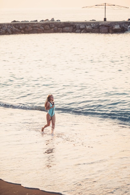 Mooi meisje in een blauwe zwembroek en hoed op een zandstrand aan zee in het zonlicht van de zonsondergang