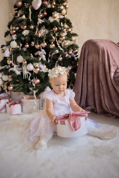 Mooi meisje in een feestelijke witte jurk, zittend op een deken in de buurt van de kerstboom opent met een cadeau