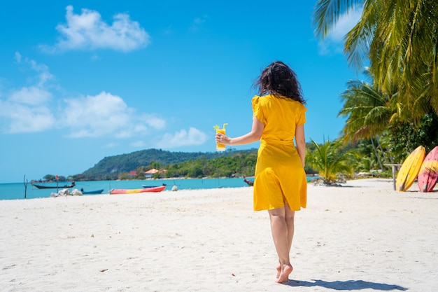 Mooi meisje in een gele jurk drinkt verse mango op het strand van een paradijselijk eiland. Perfecte vakantie.