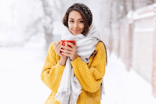 Mooi meisje in een gele trui en een witte sjaal staande met een rode mok op een besneeuwde straat