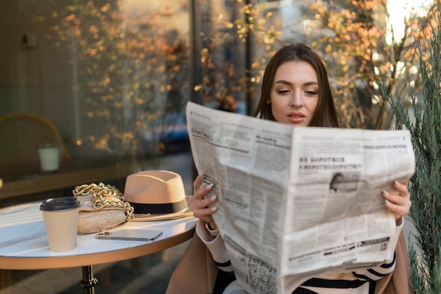 Mooi meisje in een jas leest een krant en glimlacht terwijl ze ontspant op een terrasje