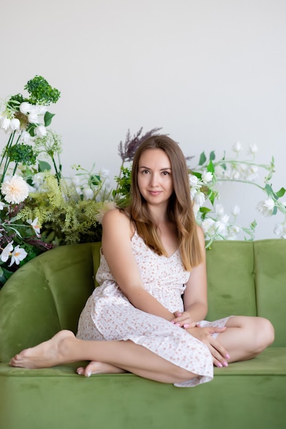 Mooi meisje in een jurk zit op een groene bank in een kamer omringd door bloemen Wegkijkend