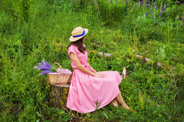 Mooi meisje in een mooie jurk zit op een boomstronk in het bos, een boeket van lupines in een mand