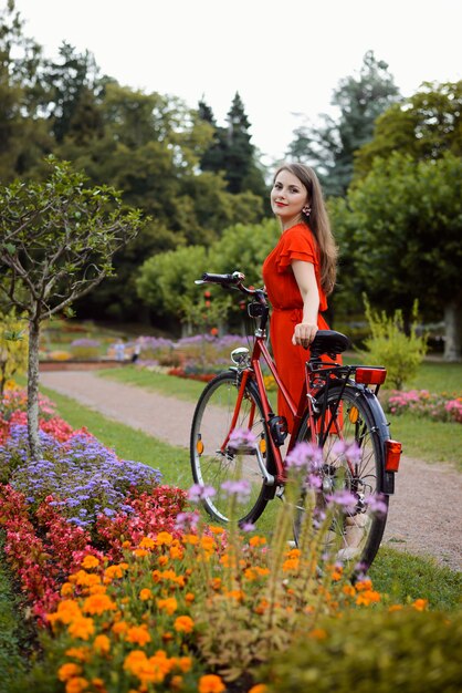 Mooi meisje in een rode jurk in de buurt van rode retro fiets in het park in de buurt van bloemen