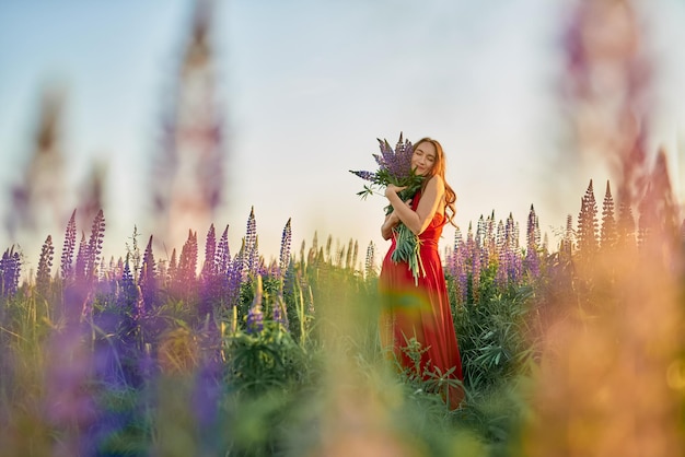 Mooi meisje in een rode jurk met een boeket lupinebloemen in het veld