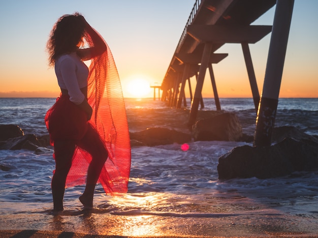 Mooi meisje in een rode jurk op het strand bij zonsopgang