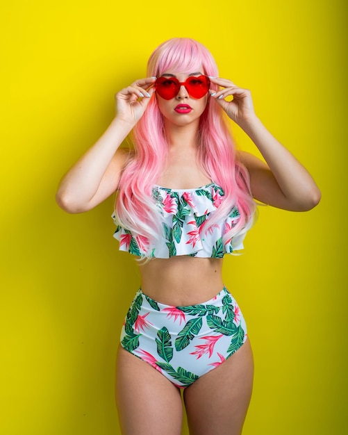 Mooi meisje in een roze pruik en gekleurde bikini poseren op een gele achtergrond Vrouw met kunstmatig lang haar en roze bril in de vorm van harten