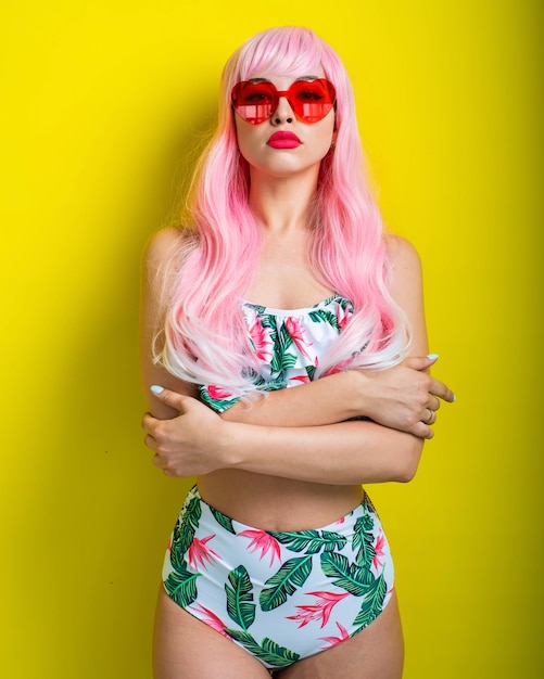 Mooi meisje in een roze pruik en gekleurde bikini poseren op een gele achtergrond Vrouw met kunstmatig lang haar en roze bril in de vorm van harten