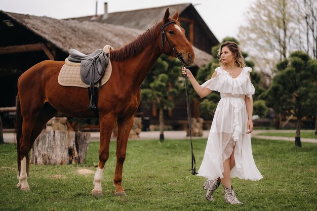 Mooi meisje in een witte zomerjurk naast een paard op een oude ranch