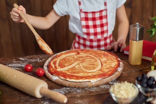 Mooi meisje in geruite schort kokende pizza, het verspreiden van saus met lepel in hartvorm