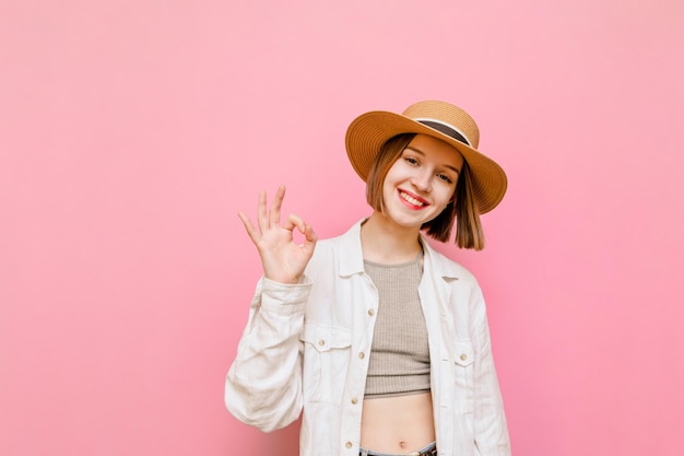 Mooi meisje in hoed en lichte zomerkleren die gebaar OK tonen en naar camera kijken geïsoleerd op roze achtergrond en knipogen Aantrekkelijke dame toerist toont OK gebaar met een glimlach op gezicht verticaal