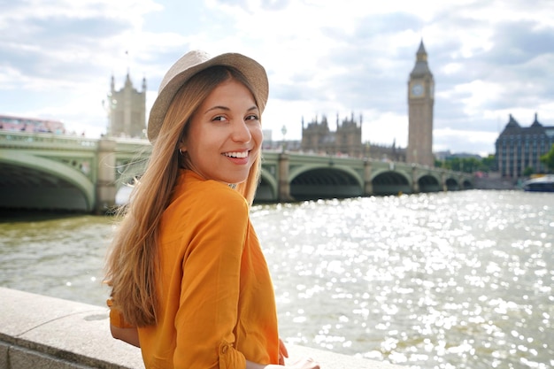 Mooi meisje in londen uk portret van jonge stijlvolle vrouw in oranje shirt en hoed camera kijken met westminster bridge op de achtergrond