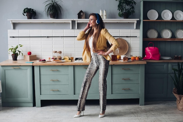 Mooi meisje in modieuze kleding krijgen plezier op keuken Gelukkig brunette model met lang haar luisteren naar muziek en dansen Stijlvolle vrouw in gele jas en blauwe koptelefoon zingen