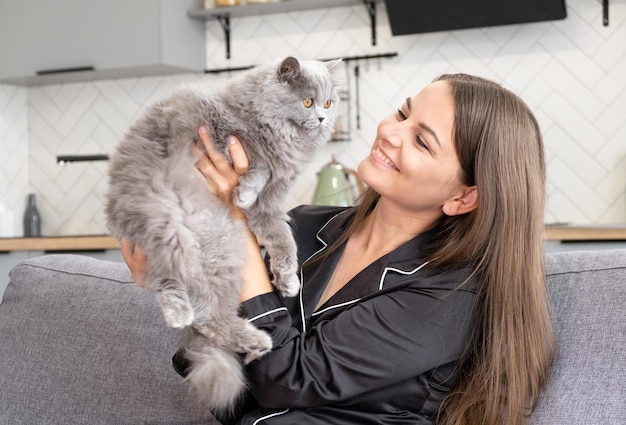 Mooi meisje in pyjama houdt haar geliefde huisdier in haar armen Britse kat in de handen van een volbloed Britse kat meisje zit op de bank in een mooie gezellige kamer