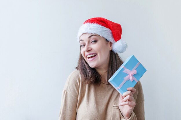 Mooi meisje in rode kerstman hoed met blauwe geschenkdoos in de hand geïsoleerd op een witte achtergrond. Jong vrouwenportret, ware emoties. Gelukkig Kerstmis en Nieuwjaar vakantie concept.