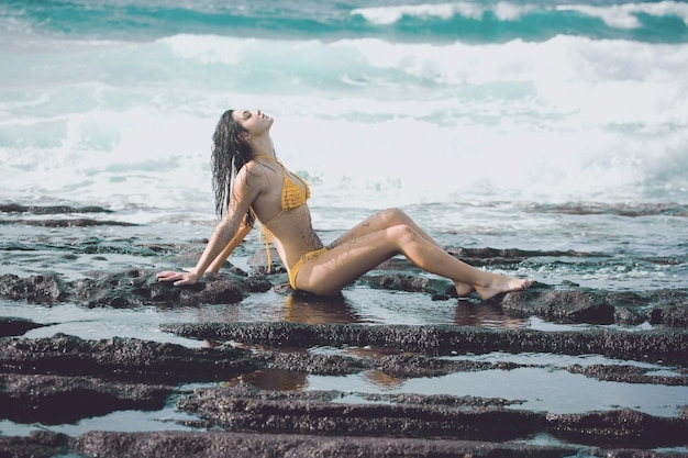 Mooi meisje in sexy gele zwembroek zittend op rotsachtig strand