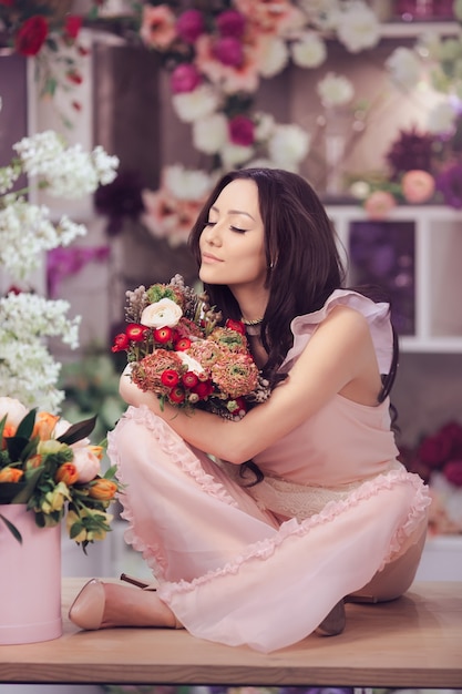 Mooi meisje in tedere roze jurk met boeket bloemen pioenrozen in handen zittend op tafel
