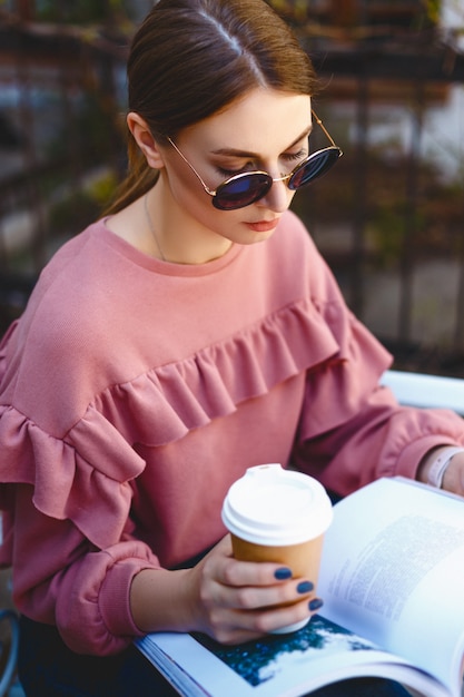 Mooi meisje in zonnebril en een roze trui zit op een bankje op een warme lentedag en houdt een kopje koffie en een tijdschrift