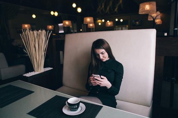 Mooi meisje kijkt naar haar telefoon, zittend op de bank in een café