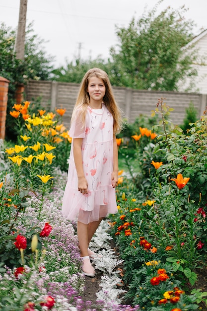 mooi meisje met blauwe ogen en lang blond haar. klein meisje in een roze flamingo jurk. meisje in de bloementuin. zomer heldere, emotionele foto. grote, dikke, lichte bloementuin bij het huis.