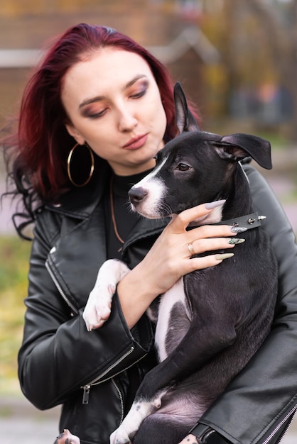 Mooi meisje met bordeauxrood haar en in een elegante jurk met een schattige puppy Houdt de hond vast Hond mix Staffordshire Terrier en Pit Bull Terrier