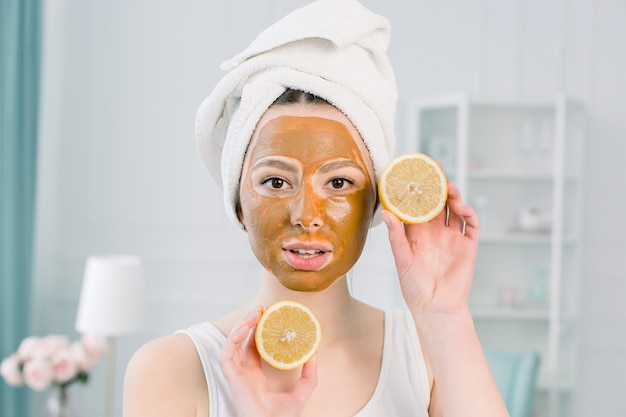 Mooi meisje met bruin gezichtsmasker die een plakje citroen houden dichtbij haar gezicht en het glimlachen. Foto van meisje dat kuuroordbehandelingen ontvangt. Schoonheid en huidverzorging concept