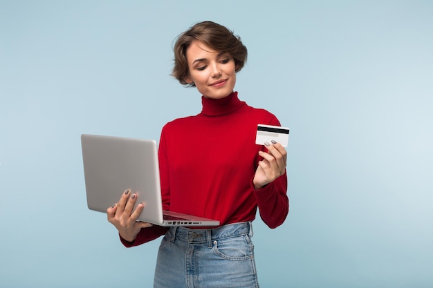 Mooi meisje met donker kort haar in rode trui en spijkerbroek die laptop in de hand houdt terwijl ze dromerig op creditcard over blauwe achtergrond kijkt